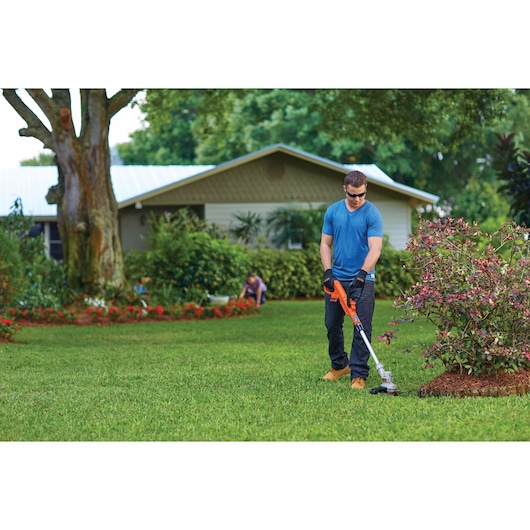 Person using BLACKDECKER LST300 string trimmer to edge grass in a garden with shrubs and a house in the background.