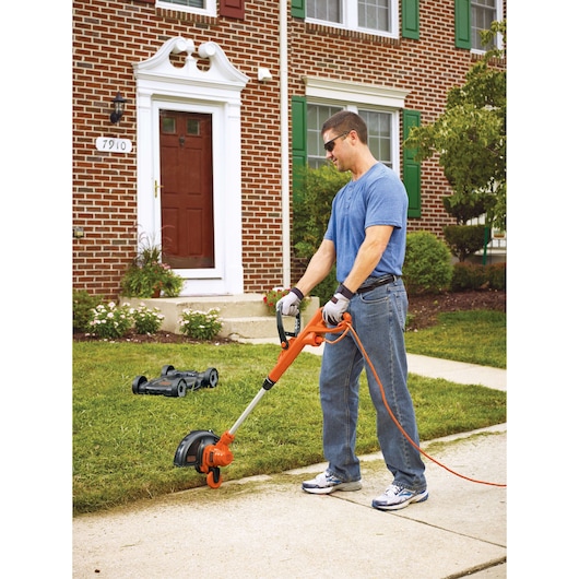 Photo of a person using a BLACKDECKER MTE912 electric lawn trimmer on a sidewalk edge in front of a brick house.