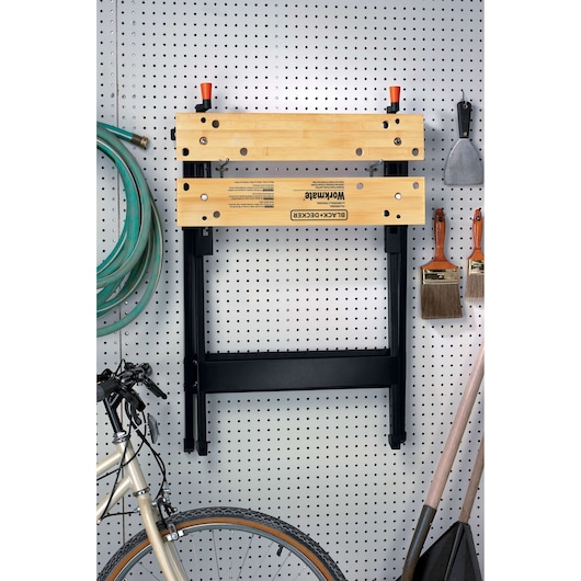 BLACK+DECKER WM125 portable workbench with wood top, folded and hanging on a pegboard wall in a garage.